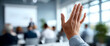 © perfectlab - Close-up of raised hand in business meeting with blurred audience and presenter in modern conference room setting