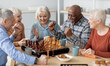 © Prostock-studio - Emotional multiethnic group of senior people playing chess, happy elderly men and women in casual outfits sitting around table with table games, chatting, having fun at sanatorium