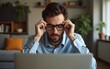 © William - Tired freelancer works at home. Frustrated young man in shirt, with laptop at remote job, takes off glasses, rubs eyes, suffering from dryness and overwork, sits at computer in living room interior