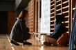 © TrueFrame Collective - Caucasian young adult woman crouching and talking to seated teenager boy with curly hair and hoodie in gymnasium setting, both engaging in conversation near wooden wall bars