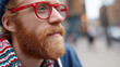 © GrayFrog - Close up on a man with red beard and stylish red glasses gazes thoughtfully against blurred urban background.