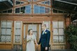 © Raivo - Bride and groom holding hands in front of rustic wooden building, showcasing love and commitment, surrounded by nature, capturing a romantic wedding moment with vintage charm