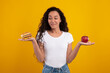 © Prostock-studio - A cheerful woman with curly hair stands against a bright yellow background, holding a red apple in one hand and a burger in the other. She looks playfully torn between healthy and unhealthy choices.