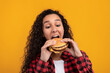 © Prostock-studio - A cheerful Latin woman bites a large hamburger in a studio setting. She looks excited while posing against a bright orange background. This scene captures a moment of enjoyment with food.