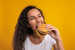 © Prostock-studio - A woman with curly hair smiles as she prepares to bite into a large burger. She is in a studio with a bright yellow background, showcasing her love for tasty food and fun moments.