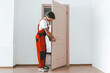 © Prostock-studio - A construction worker in red overalls is fitting a wooden door in a bright, modern room. The space has light-colored walls and a clean, open layout with natural light.