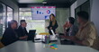 © Marco - Businesswoman explains data chart to engaged team during office presentation while group of professionals seated around conference table watch screen