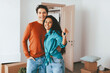 © Prostock-studio - A happy couple stands together, smiling while holding up a set of house keys. They are in a room with moving boxes, indicating they just moved into a new home.