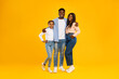 © Prostock-studio - A joyful family stands together in front of a vibrant yellow-orange wall. The husband hugs his wife while their smiling daughter poses playfully beside them. This scene radiates love and happiness.