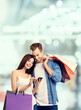 © vgstudio - Vertical banner image with free ad text space - sale, price discount, consumer credit concept - happy couple hold shopping bags, gazing at smartphone. Man, woman stand against blurred mall background