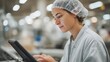 © CYBERPINK - Young woman wearing a white hairnet on her head and a grey lab coat. she is standing in a factory or workshop, looking intently at a black tablet computer in her hands.