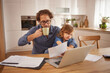 © Stockphotodirectors - A father drinks coffee while seated at a kitchen table, focused on his laptop. His daughter explores papers next to him, enjoying quality family time in a cozy home environment.