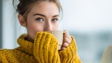 Beautiful young woman with striking blue eyes is enjoying a warm drink. Dressed in a cozy yellow sweater, she sits in a bright, inviting cafe, relishing the peaceful atmosphere and her beverage