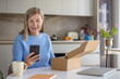 © Liubomir - Senior woman smiling, holding smartphone, and looking at a cardboard delivery box, excited about her online shopping purchase in a modern home kitchen