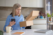© Liubomir - Woman feeling disappointed and confused after unboxing an online shopping package, holding a smartphone, discovering a wrong order or missing item while sitting at her kitchen table