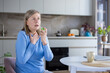 © Liubomir - Senior woman sitting at kitchen table spraying throat with relief medicine for sore throat and cough, showing home healthcare and illness treatment in casual domestic setting