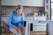 © Liubomir - Senior woman sitting in her kitchen gripping her painful knee, showing discomfort from age-related joint pain and arthritis affecting everyday home life and mobility