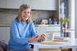 © Liubomir - Happy senior woman sitting at a table in her home kitchen, smiling and engaging in an online video call using a digital tablet, connecting with family or friends