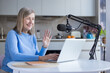 © Liubomir - Senior woman smiling and waving during a video call or recording session, actively creating digital content and connecting remotely using a microphone and laptop in her modern kitchen
