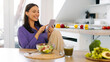 © Home-stock - Positive young lady eating fresh salad and using cellphone, sitting at table in kitchen while having healthy dinner, copy space