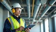 © Maryna - Asian engineer man wears safety gear. He inspects industrial plumbing pipes using tablet. Worker checks building system with tech. He reviews plans and checks quality. Focused man works on project.