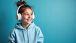 © Maryna - Smiling teen girl wears headphones. Young female with earphones listens music on blue background. Happy woman in hoodie enjoys audio. Joyful student listens playlist or song.