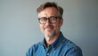 © Maryna - Middle aged man with glasses smiles at camera. Wears blue denim shirt. Confident male adult looks directly in studio. Mature guy with short beard poses for headshot on grey background. Person looks