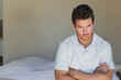 © wavebreak3 - Man sitting on bed edge in bedroom wearing light polo, crossing arms, copy space