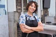 © wavebreak3 - Mature female stylist standing arms crossed inside salon wearing black apron with scissors comb