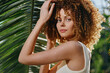 © SHOTPRIME STUDIO - Curly-haired woman posing outdoors in white top near tropical palm leaves with natural sunlight and green blurred background under warm summer conditions.