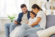 © Studio Romantic - Happy family parents and child at home. Smiling daughter on couch with mother and father using tablet together. Warm living room shows trust, care, and unity. Technology and loving family time.