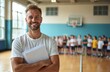 © Vadym - Smiling male coach holds papers in school gymnasium. Blurred children in sports attire stand ready. Indoor physical education lesson begins. Healthy active lifestyle motivation.