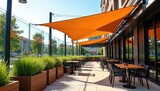 Modern cafe patio features orange shade sails over tables, chairs. Rich green grass planters line tiled floor. Sleek glass wall reflects trees, buildings. Outdoor dining ambiance is bright, airy.