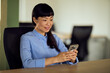 © bnenin - Asian Woman in Blue Shirt Using Smartphone in Modern Office, Calm and Focused at Desk