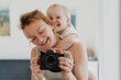 © nataliaderiabina - Smiling woman holds camera while laughing child hugs her from behind, capturing joyful moment together at home. Happy mother with daughter spending quality time together taking selfies on camera