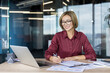 © Liubomir - Young happy professional woman wearing glasses and a maroon shirt sitting at her office desk, writing on paperwork and using a laptop, representing productivity and modern work environment