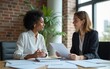 © happy - Two business women sit at desk discuss project details, diverse female colleagues met in office, share opinion, working on collaborative task, sales manager makes commercial offer to company client