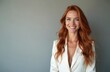 © Viktor - Confident woman smiles happily at camera. Long red hair, green eyes. Female manager poses in white suit. Successful professional lady stands against plain grey background. Friendly expression