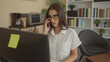 © Krakenimages.com - Woman wearing glasses holds phone to ear while typing on keyboard at desk in office building with bookshelves behind; concentration.