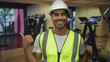 © Krakenimages.com - Man wearing hardhat and neon vest smiles and gives thumbs up gesture in gym building; confidence safety.