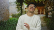 © Krakenimages.com - Man wearing white sweater and glasses laughs and waves hand at stone building entrance with garden pathway; joy.