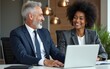 © lan - Happy professional mature Latin businessman and businesswoman executive colleagues wearing suits sitting at table having partnership business discussion using laptop at corporate meeting in office.