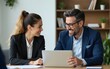 © lan - Happy professional mature Latin businessman and businesswoman executive colleagues wearing suits sitting at table having partnership business discussion using laptop at corporate meeting in office.