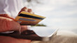 © wkproduction - A digital nomad's hand holds a credit card near a laptop, symbolizing remote work and financial transactions. The blurred natural background suggests a serene, portable workspace.
