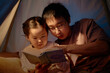 © Seventyfour - Asian man and child reading book together inside home tent, father guiding daughter through story, both focused on pages, spending evening engaged in family activity