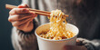 © Bonsales - Hands picking up steaming ramen noodles from a disposable cup container with wooden chopsticks, representing convenience food