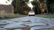 © Canary4stock - Ancient Roman road made of large cobblestones with a modern car driving away, blending historical infrastructure with contemporary travel and pine trees along the path