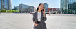 © insta_photos - Young Asian successful businesswoman leader wearing suit standing in big city talking on mobile phone. Smiling woman making business call on cell walking on busy downtown street outdoors