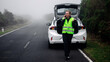 © wedmoments.stock - A woman wearing a reflective vest talks on a mobile phone next to a white car with an open trunk on a foggy road, showing emergency help and support.