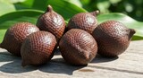 Six brown, scaly snake fruits sit on weathered wood, backlit by green leaves